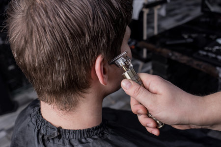 The hand of a barber or hairdresser cuts the dark long hair of the guy's client on his temple....