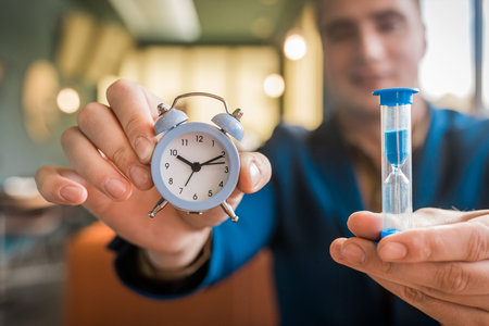 The hand of a male businessman holds a clock alarm time and an hourglass, close-up.の写真素材