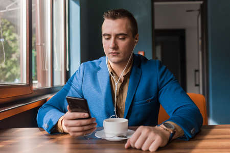 A stylish guy businessman of European appearance in a jacket and shirt sits at a table in a cafe on a coffee break, holds a smartphone or mobile phone in his hand.の写真素材