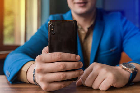 Close-up, businessman guy holds a black smartphone or mobile phone in his hand and takes a selfie or talks on a video link in a cafe.の写真素材