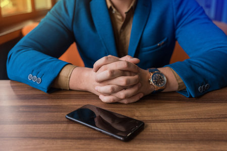 A businessman in a blue suit, jacket with a watch in his hands sits idly by next to a mobile phone...