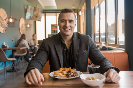 A young satisfied guy portrait of a businessman in a jacket sits at a table in a cafe on a lunch...