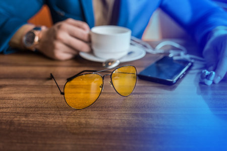 Sunglasses, mobile phone or smartphone and a mug of coffee on the table in a cafe with the hand of a businessman.の写真素材