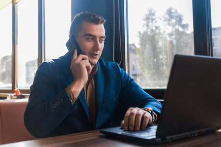 A business man surprised businessman in an attractive European-looking suit works in a laptop, talks on a cell or mobile phone, sitting at a table in a cafe by the window.の写真素材