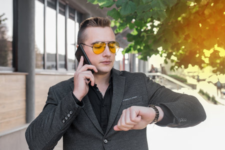 A young serious guy, businessman of European appearance portrait in sunglasses talks on a mobile phone or smartphone and looks at the time on a hand watch on the street outdoor.の写真素材