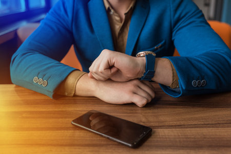 A businessman in a blue jacket looks at the time on a hand watch, opposite lies on a wooden table of...