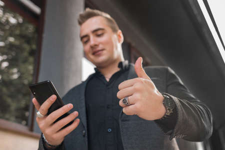 A young positive man in a gray jacket and black shirt holds a mobile phone or smartphone and points the class with his finger up on the street outdoor, close-up.の写真素材