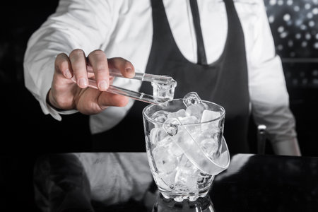 The hand of a professional bartender holds plastic ice cube tongs next to a cold ice dish.の写真素材