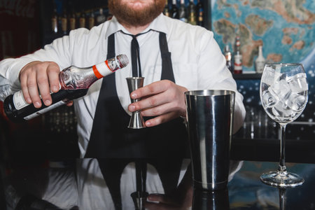 The hands of a professional bartender pour red syrup into a measuring glass of jigger, next to a metal tool for preparing and stirring alcoholic cocktails of shaker drinks and glasses with ice cubes.の写真素材