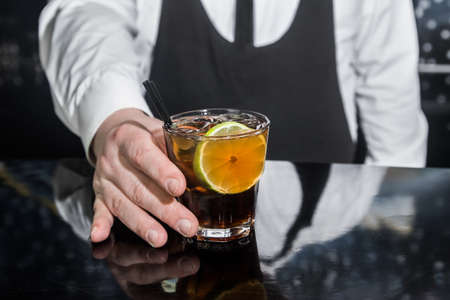 The hand of a professional bartender serves an alcoholic beverage cocktail whiskey with cola and a piece of lime on the bar counter, close-up.の写真素材