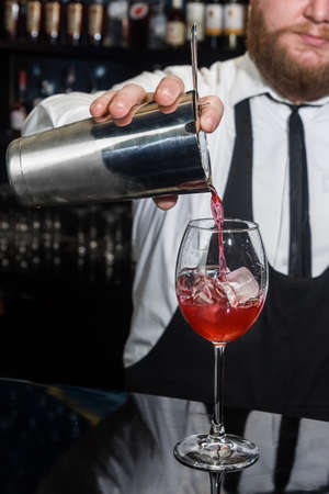Professional bartender with a beard pours liquid into a glass of ice cubes from a tool for mixing and preparing alcoholic beverages cocktails metal shaker.の写真素材