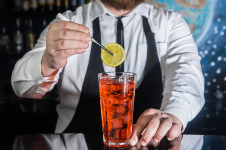 A professional bartender puts a slice of lemon in a red chilled alcoholic cocktail with bar tweezers in glass at a nightclub counter.の写真素材