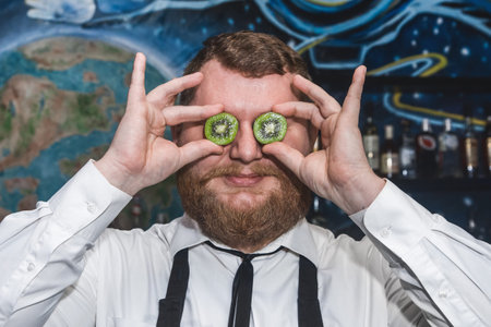 Adult funny bearded positive man professional bartender holds halves of sliced kiwi in his hands at the face.の写真素材