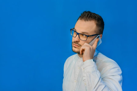 Young attractive businessman in a white shirt and glasses speaks on the phone and smiles on a blue...