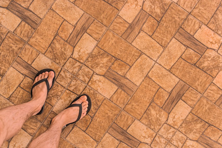 Legs of a man in black flip flops stand on a brown tiled floor with an abstract stone pattern...