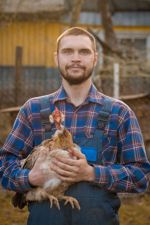 Farmer smiling positive European appearance male Caucasian rural portrait on countryside with a beard, shirt and overalls with a white-necked chicken in his arms outdoors.の写真素材