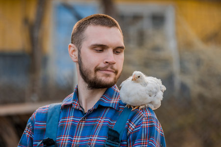 A farmer of European Caucasian appearance a smiling man with a beard portrait looks at the dwarf white chicken on his shoulder, close up against the backdrop of the countryside.の写真素材