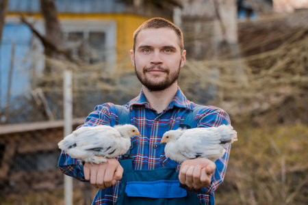 A satisfied farmer a man with a beard, in overalls and a shirt, holds two white dwarf chickens in his arms against the backdrop of a countryside.の写真素材