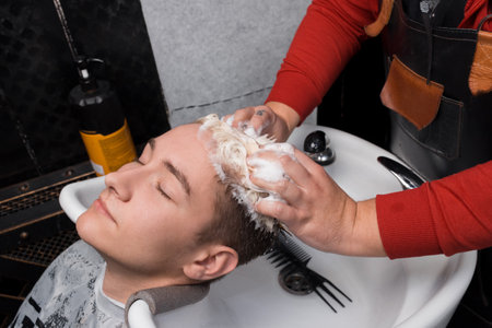 The hands of a professional barber lathered with foam the hair of a client's head of a guy of European, appearance in a hairdresser before a haircut.の写真素材