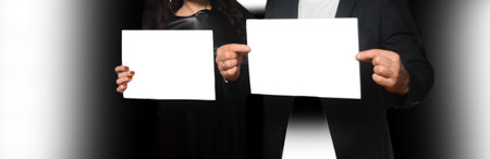 Two people, a man and a woman, holding a sheet of A4 paper in their hands, an empty space for text and design white blank on a dark background.の写真素材