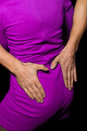 The hands of an adult man lie on the waist of a woman in a pink dress while dancing on a dark...