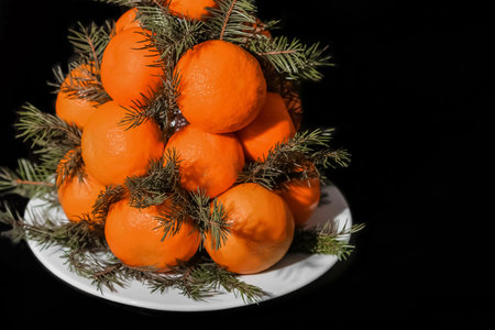 A group of piles of many tangerines decorated with spruce branches on a white plate on a black...