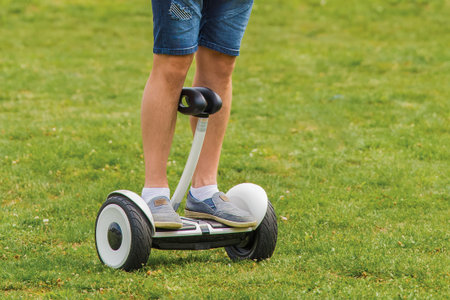 The legs of an adult man stand on a hoverboard against the background of grass in the park.