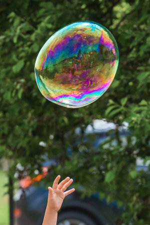 A child's hand reaches up to a large colored soap bubble on the blurred green background of the...