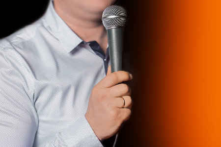 Person's hand with a ring on his finger holding a professional instrument karaoke equipment microphone for singing on a black orange background close-up.の写真素材