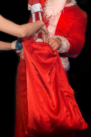 The hands of Santa Claus in a New Year's Santa costume climb and and Snow Maiden, into a red bag with gifts close-up on a dark background.の写真素材