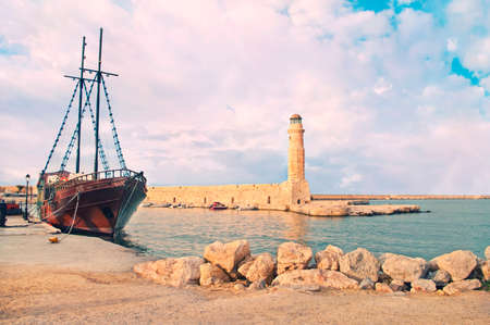 cross-processed image of old Venetian Lighthouse with old-style wooden ship moored in dock in warm sunset colours, Rethymno, Crete, Greeceの写真素材