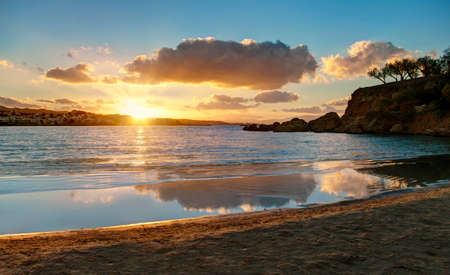 beautiful sunset over small city from sandy beach with small rocky islet on right, Crete, Greeceの写真素材