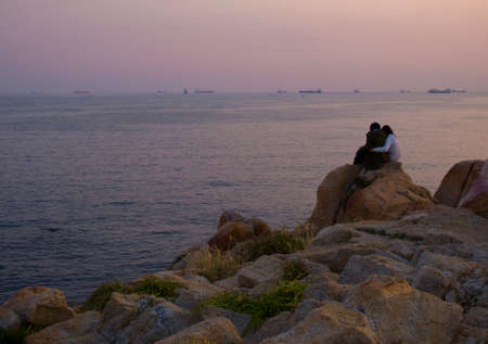 korean couple sitting on rocks watching sunset over seaの写真素材