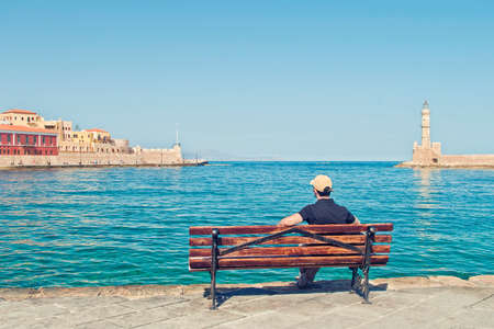 young caucasian male tourist wearing black t-shirt and baseball cap sitting alone on bench and looking at old part of Chania and venetian lighthouse on sunny summer day, Crete, Greeceのeditorial素材