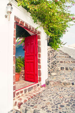 red open door of a house with blooming plant over it on warm spring day with boulder stairs in Oia, Santorini or Thira, Greeceの写真素材