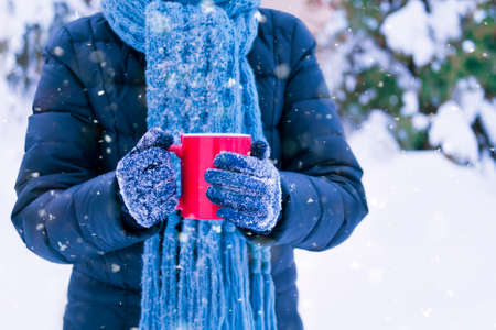 young woman in blue jacket wearing blue knitted scarf standing outside on snowy winter day holding red cup with warm drinkの写真素材