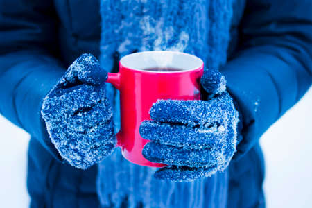 female hands in gloves holding red cup with steaming hot dark drink outside on winter dayの写真素材