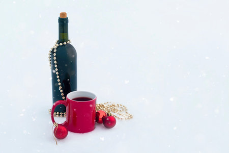 bottle of wine with red mug and christmas ornaments on snow with snow falling from sky