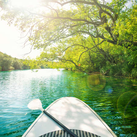 white paddle board with oar floating along clear waters of Zrmanja river surrounded by trees and greenery on sunny summer day, Zadar, Dalmatia, Croatiaの写真素材