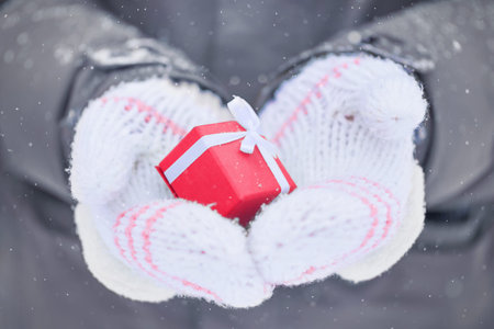 two hands in white mittens holding a small red box with a present outside on snowy winter day