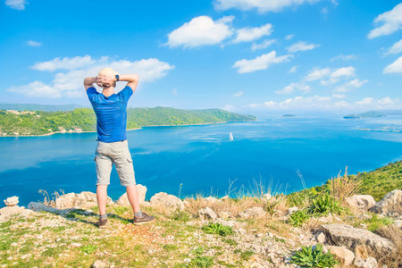 young caucasian man standing at cliff looking at sea view and islands on sunny summer day, Dugi...