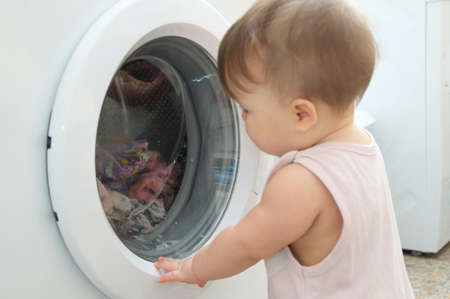 Baby near washing machine looking at camera,  having fun and playing with washerの写真素材