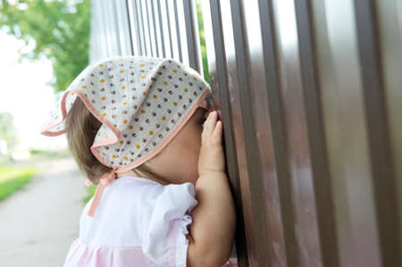 Baby girl peeping through hole in fence. Child looking on somethingの写真素材