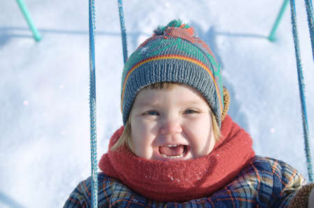baby in winter clothes playing on swing outdoor. happy smiling cute child in cap,scarf among the snowの写真素材
