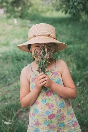 Young beautiful Asian brown haired woman in a hat smiles behind a bouquet of wild flowers. Green grass backgroundの写真素材