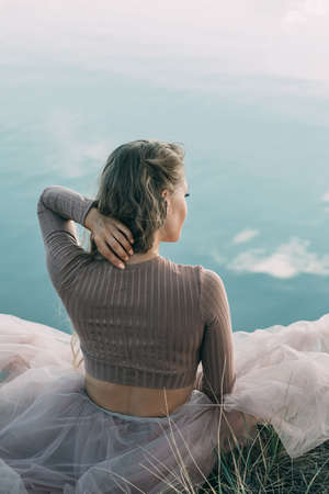 Outdoor recreation. A woman sits on the edge of a cliff near the lake touches her hair with her handの写真素材