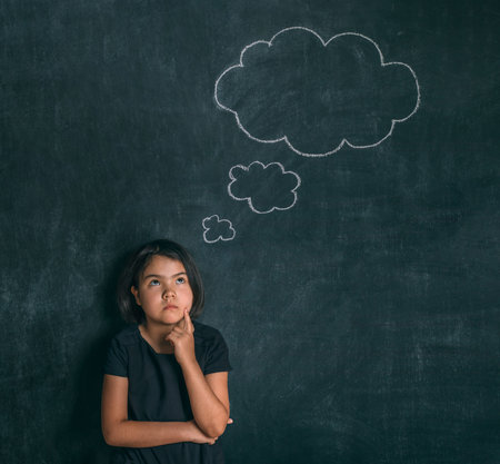 Back to school. Serious little girl thinking touching her head with a finger near blackboard. Child...