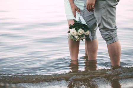 The view of the newlyweds legs standing on the beach. The view of the wedding bouquet of white roses and cottonの写真素材