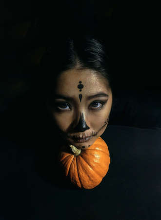 A young beautiful girl with skeleton makeup put her head on a pumpkin on a black background. The concept of the Halloween celebration. Dark photoの写真素材