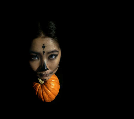 Young beautiful girl with skeleton makeup put her head on a pumpkin on a black isolated background. The concept of the Halloween celebration. Dark photoの写真素材